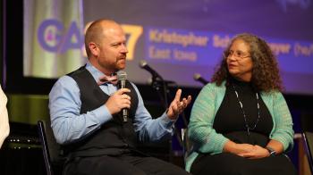 A white man with short hair in a blue button up shirt and black vest speaks into a microphone while gesturing out at an unseen audience. Beside him, a woman with gray curly hair watches him speak. 