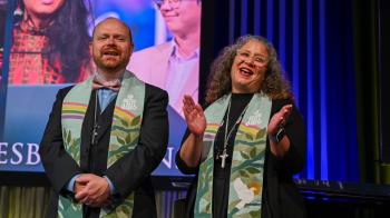 A man with a shaved head and a woman with grey and black curls stand together in stoles and robes smiling and gesturing to an unseen audience in front of them
