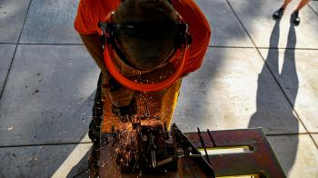 A person in an orange shirt and protective gear cuts metal, sending sparks flying, while another person stands nearby on a paved surface.