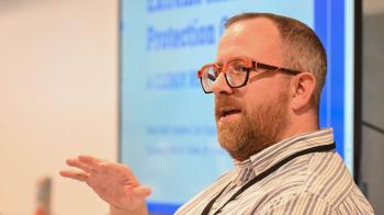 In the foreground, a white man with short hair, a beard, and glasses faces out and to his right, speaking to an audience. Behind him a screen shows a slide talking about Extreme Risk Protection Orders