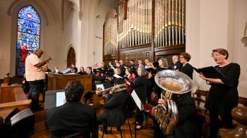 A choir and brass musicians perform in a church in front of ornate organ pipes