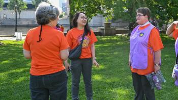 Three women in orange shirts, one with a purple vest, stand together laughing in a park.