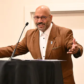 A man in glasses and a brown jacket, with arms wide, speaks at a lectern