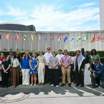 A group of 16 young adults representing at Presbyterian delegation to the UN's High Level Political Forum post together in front of UN headquarters in NYC, with flags of various nations in the background