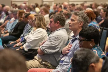 A man closes his eyes and places his hand on his chest while listening to a speaker.
