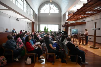 A large group of people fill a chapel with large windows at the back.