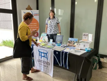 A woman stands behind a table with pamphlets while another woman leans down to read a pamphlet 