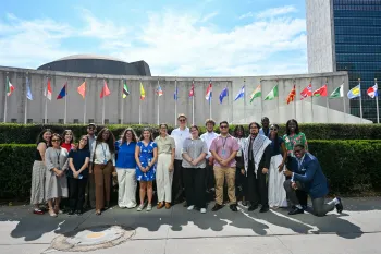 A group of 16 young adults representing at Presbyterian delegation to the UN's High Level Political Forum post together in front of UN headquarters in NYC, with flags of various nations in the background