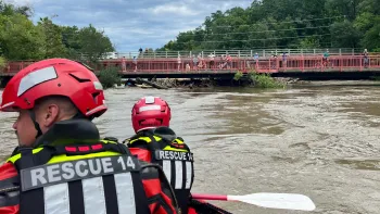 The image shows a flooded river, and two people in a boat wearing helmets and rescue gear