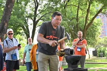 A person hammers a piece of metal on an anvil while others observe in a park setting.