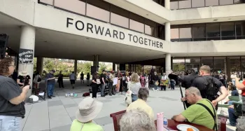 People gather, sitting and standing, as part of a vigil in front of a building that says "Forward Together."