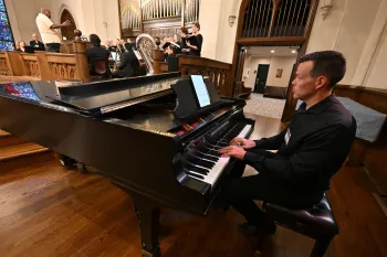 A pianist is in the foreground and a choir and brass musicians are in the background