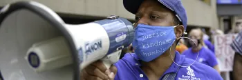 A woman carries a bullhorn in a protest while wearing a face mask that says, "justice, kindness, humbly."