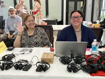 Two seated women at a table with sets of headphones look straightforward smiling 