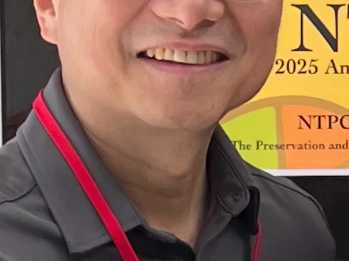 Headshot of Rev. Ralph Su wearing grey polo shirt and red lavalier with colorful yellow banner behind him