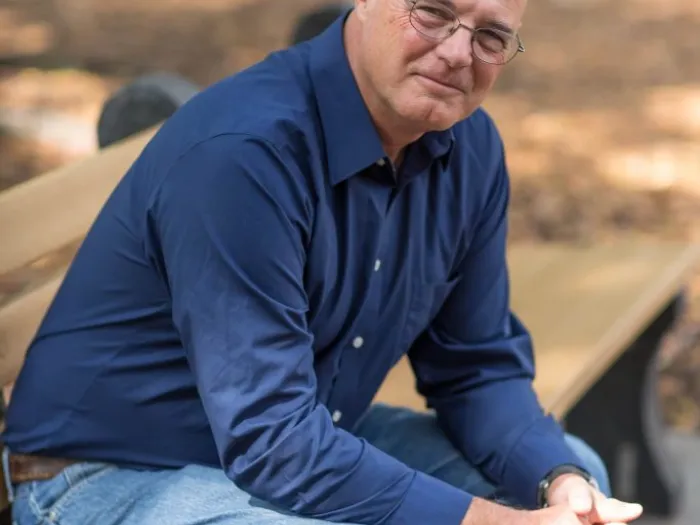 Man on bench in jeans and blue shirt with glasses, leaning over with elbows on knees