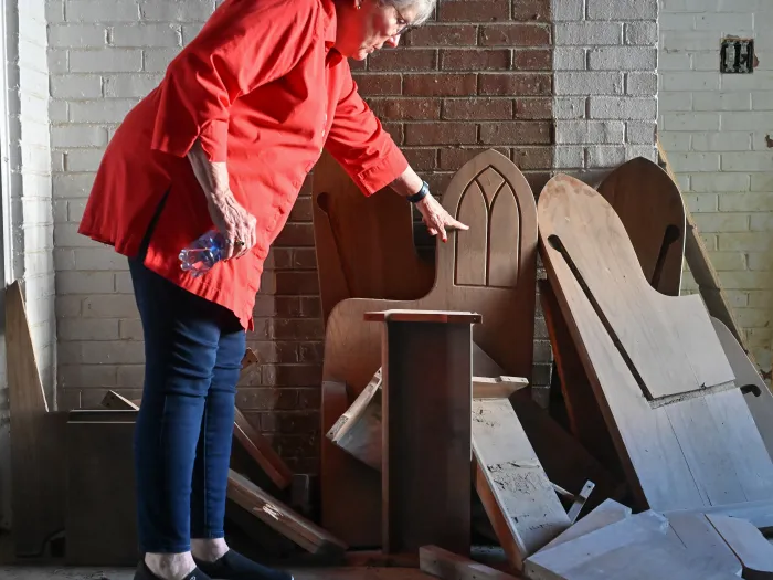 In a church basement, a woman in a red blouse points to the sides of pews that were damage and disassembled in flooding from Hurricane Helene.