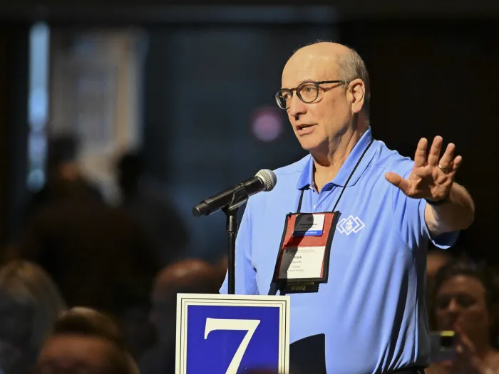 A man in a light blue polo shirt speaks at a microphone in a large meeting.