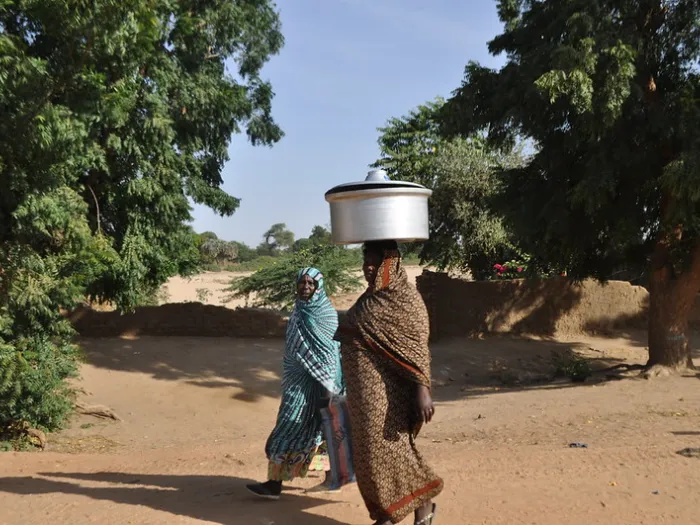 Two women walking outside, including one carrying a vessel on her head.