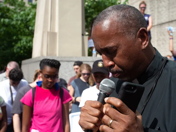 A man holding a microphone with his head bowed as people stand nearby.