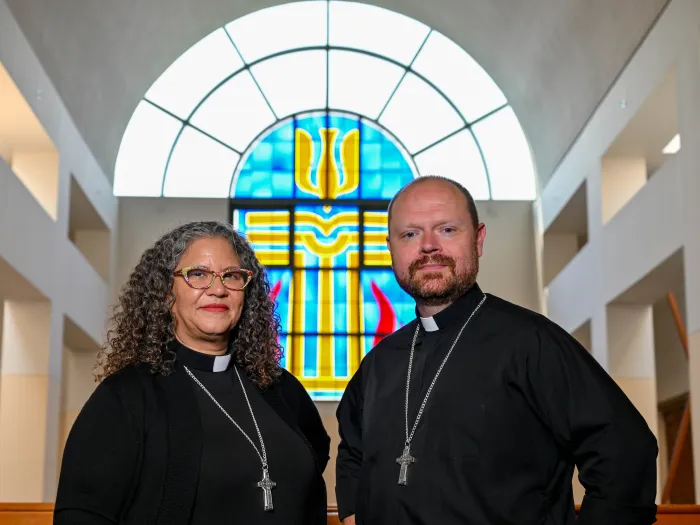 A man and a woman stand side by side in clergy collars and black shirts with silver cross necklaces. A stained glass window is behind them.