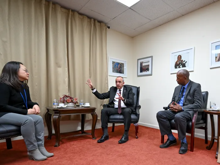A Cuban man in a business suit sits in a chair and  talks while gesturing with one hand raised. On either side of him, an Asian woman and a Black man, both in business attire, lean in to listen.