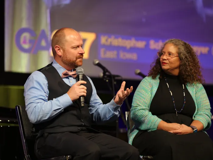 A white man with short hair in a blue button up shirt and black vest speaks into a microphone while gesturing out at an unseen audience. Beside him, a woman with gray curly hair watches him speak. 