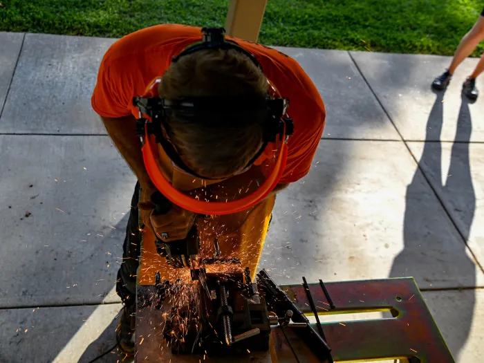 A person in an orange shirt and protective gear cuts metal, sending sparks flying, while another person stands nearby on a paved surface.