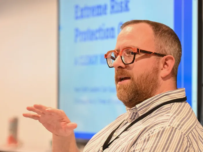 In the foreground, a white man with short hair, a beard, and glasses faces out and to his right, speaking to an audience. Behind him a screen shows a slide talking about Extreme Risk Protection Orders