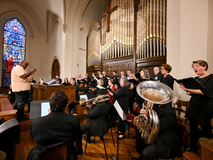 A choir and brass musicians perform in a church in front of ornate organ pipes