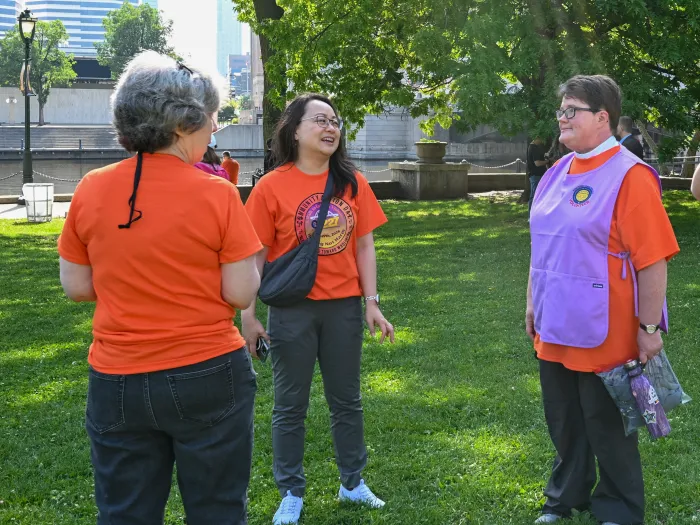 Three women in orange shirts, one with a purple vest, stand together laughing in a park.