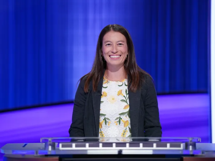 A woman in a dark blazer and white floral blouse at a game-show lectern in front of a blue curtain.