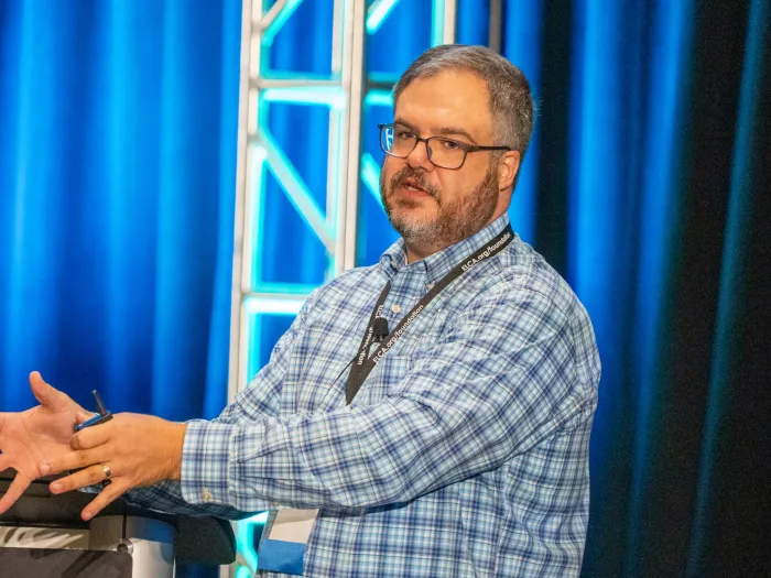 A man in a blue shirt against a blue backdrop speaks to an unseen audience.