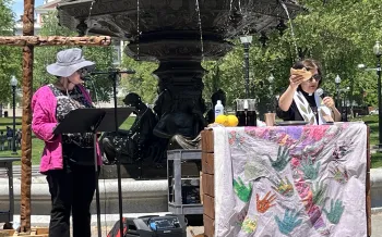 Tamar Wasoian holds a broken piece of bread above a communion table in the Boston Common. To her right, another pastor preachers from a stand.
