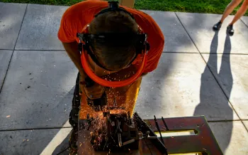 A person in an orange shirt and protective gear cuts metal, sending sparks flying, while another person stands nearby on a paved surface.
