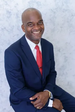 A seated man in a dark suit and red tie smiles.