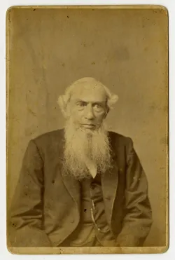 A sepia toned portrait photograph of an old white man with a long white beard that falls past the lapels of his jacket, and white hair that settles in puffs on both sides of his forehead. Reverend John Anderson, aged 80 at the time of being photographed, in 1884